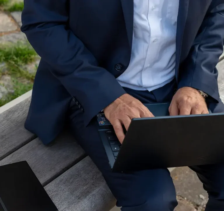 Mann in Anzug sitzt auf einer Bank und tippt auf einem Laptop.