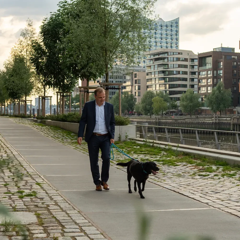 Mann in Anzug geht mit schwarzem Hund entlang einer Uferpromenade.