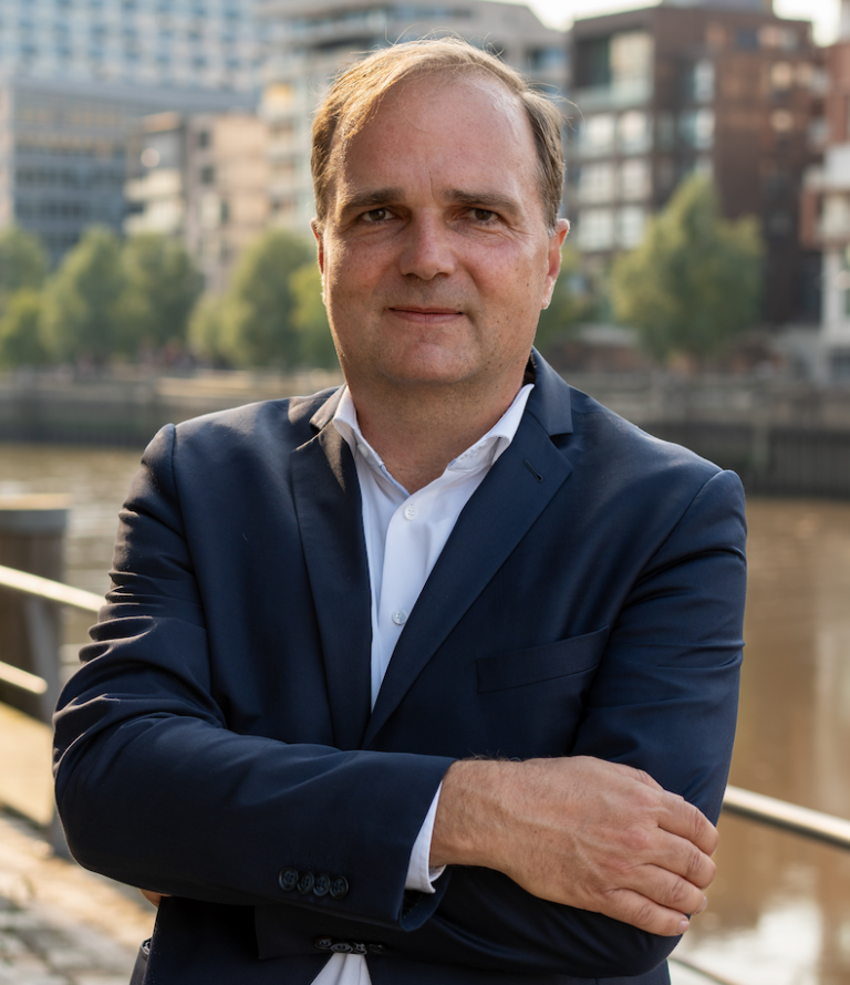 Mann in Anzug mit verschränkten Armen, Hintergrund mit Wasser und Gebäuden.